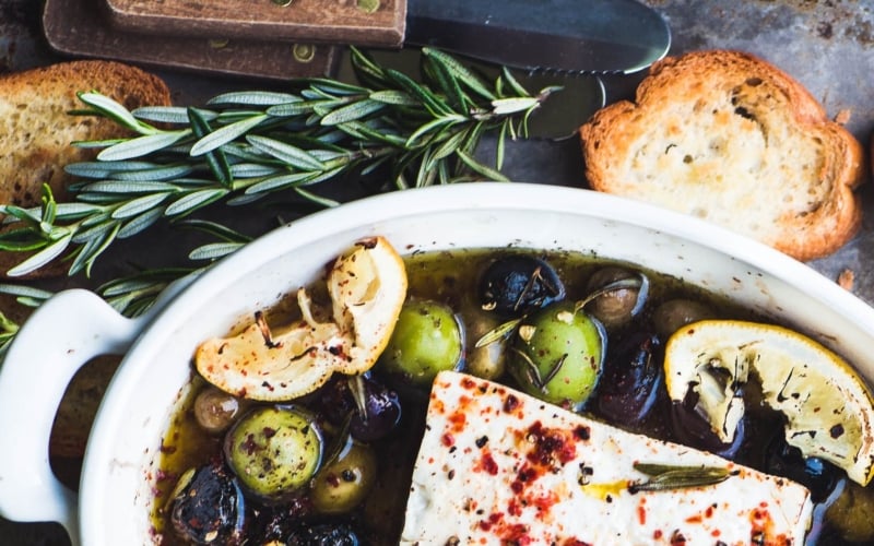 Baked feta cheese with olives, lemon, and rosemary with toasted bread on a tray with toasted bread