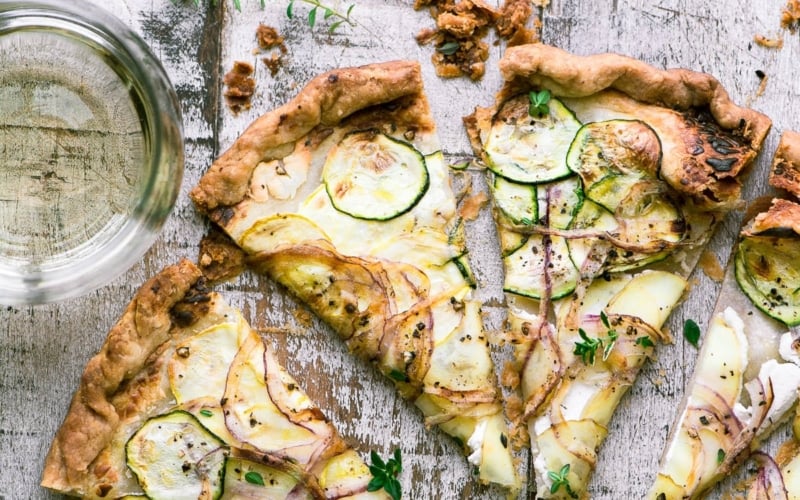 Savory goat cheese and summer squash tart sliced on a wooden table with a glass of wine