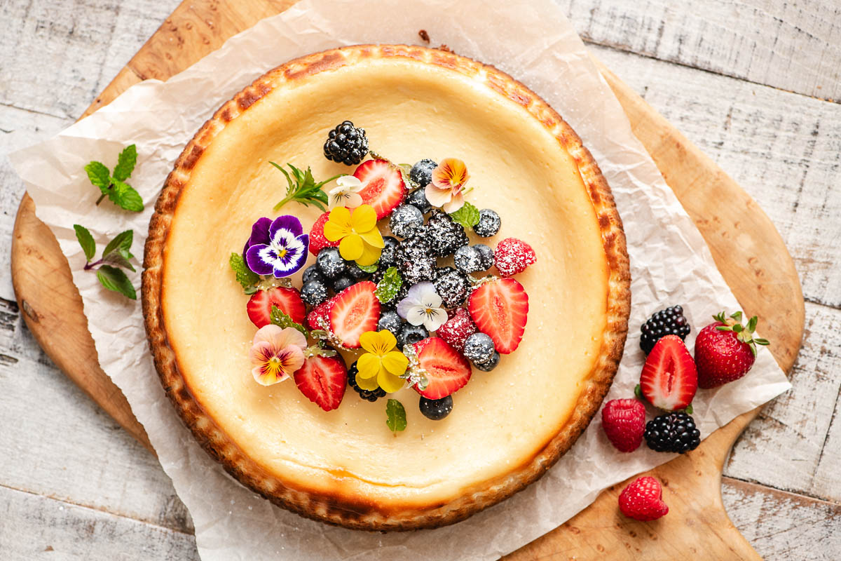 Lemon cheesecake on a wooden platter topped with fresh berries and edible flowers.