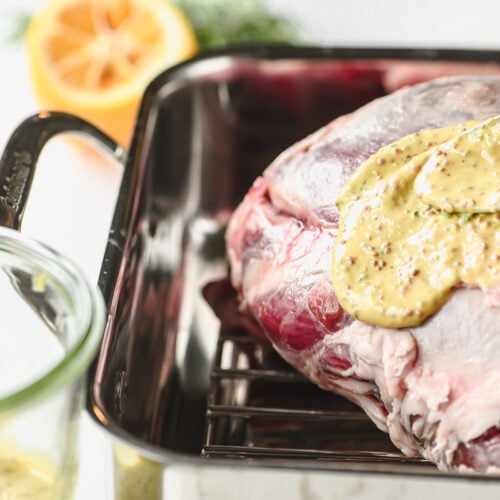 Spooning mustard coating over leg of lamb.