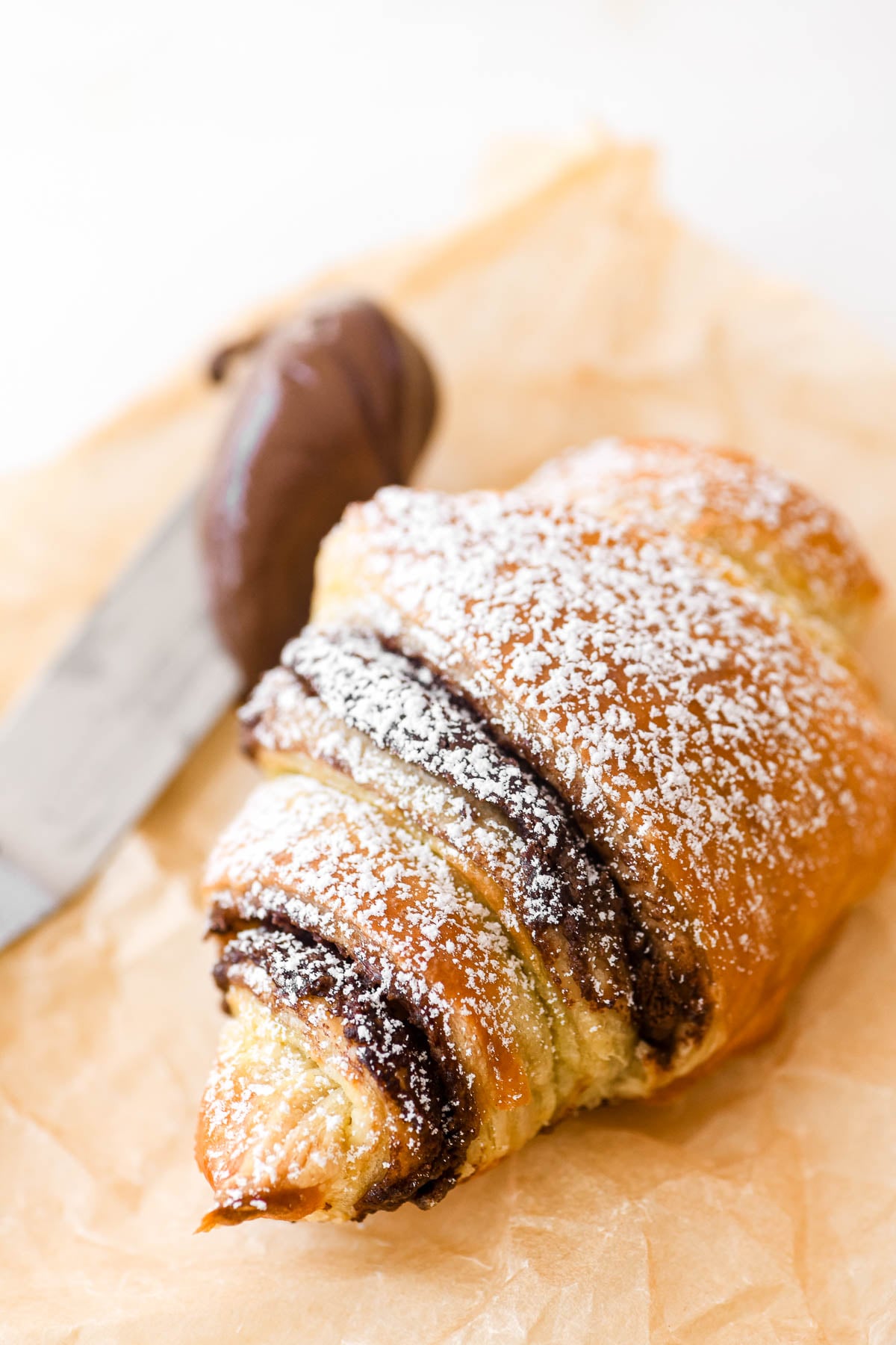 Nutella puff pastry croissant on parchment paper with knife slathered with Nutella chocolate hazelnut spread.