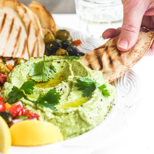 Scooping cilantro lemon hummus with a grilled pita chip.