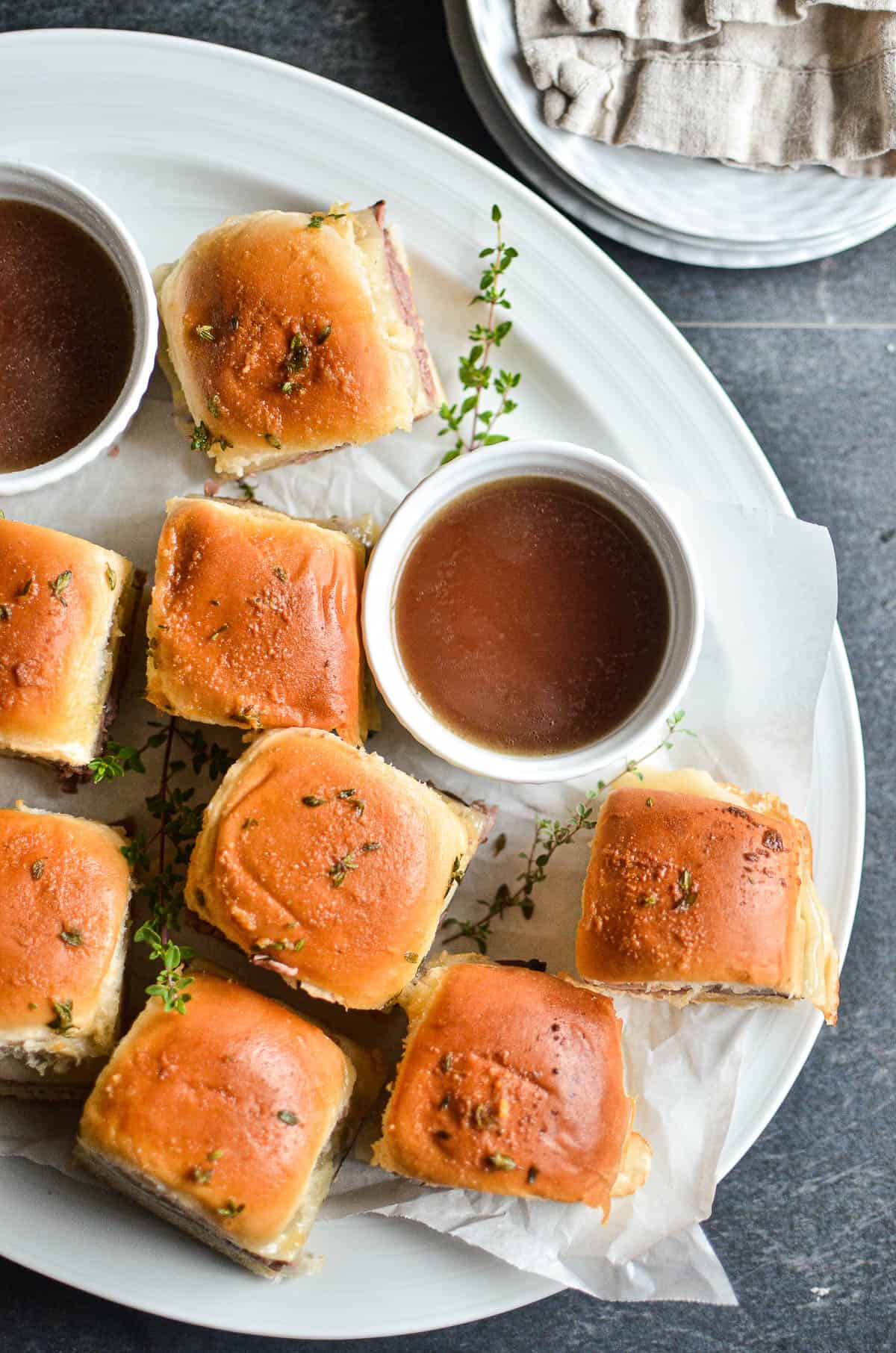 French dip sliders with ramekins of broth on a white platter.