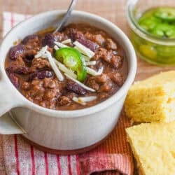 a mug of spiced chili topped with cheese and pickled jalapeños