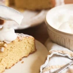 spooning whipped cream onto a slice of almond olive oil cake.