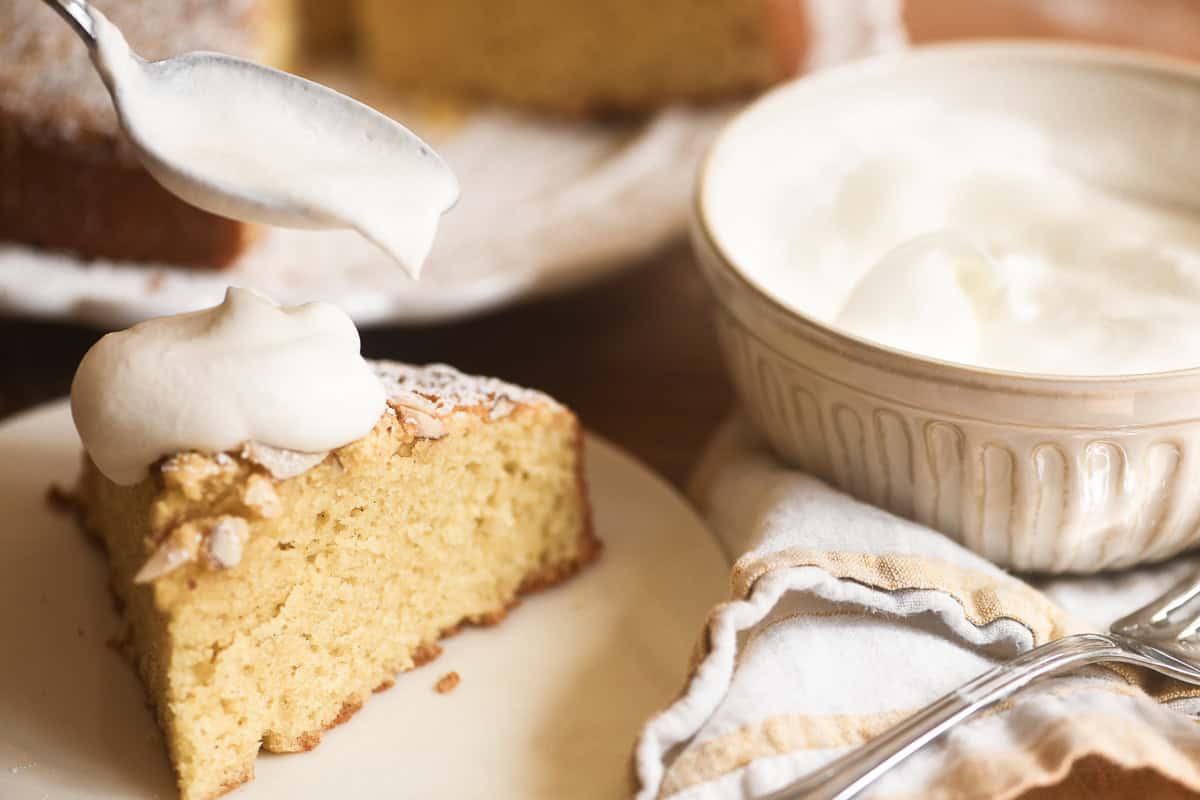spooning whipped cream onto a slice of almond olive oil cake.