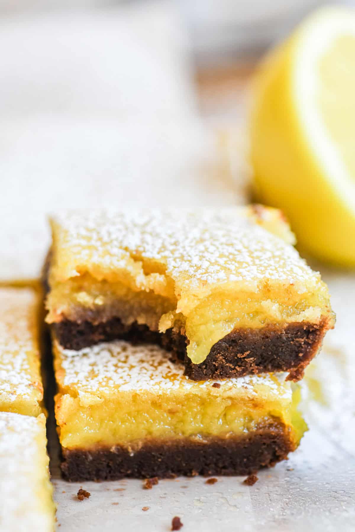 a stack of bright yellow black bottom lemon bars, with cut lemon in background.