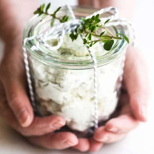 Holding a small jar of homemade Boursin style cheese tied with a ribbon for gifting.