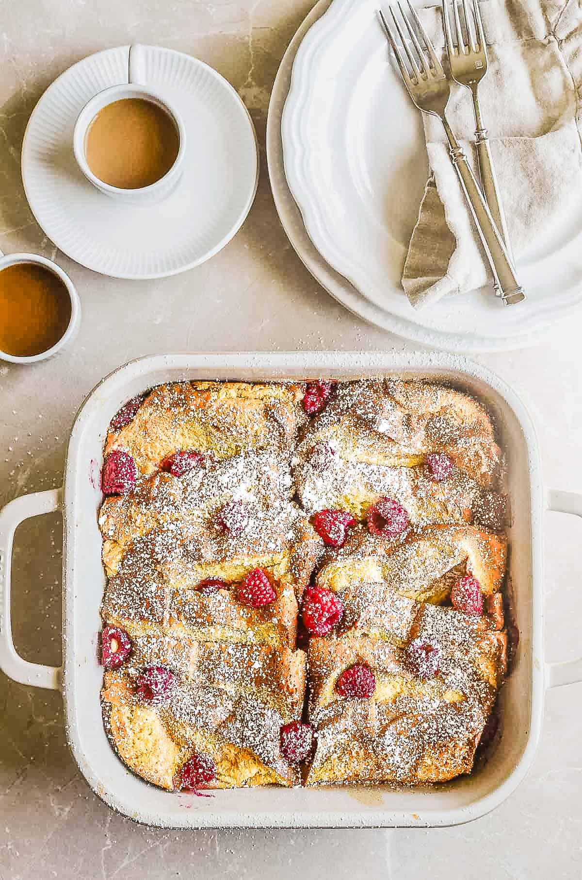 A pan of raspberry baked French toast with coffee cups and plates.
