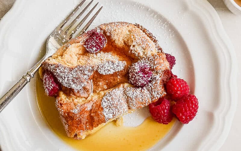 A piece of raspberry baked French toast on a plate with syrup and fresh raspberries.