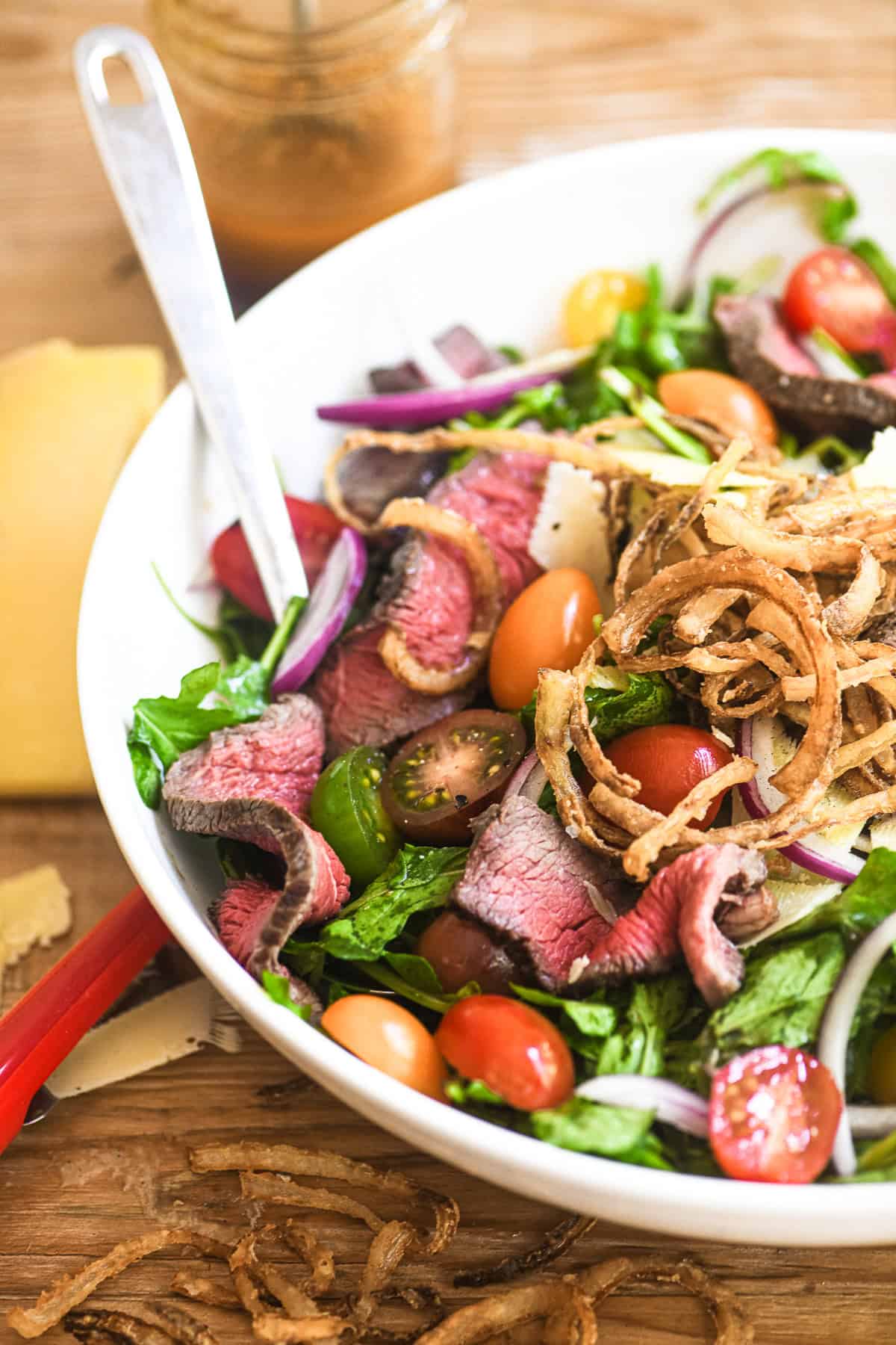 steak salad in a white bowl, topped with onion strings and shavings of Manchego cheese.