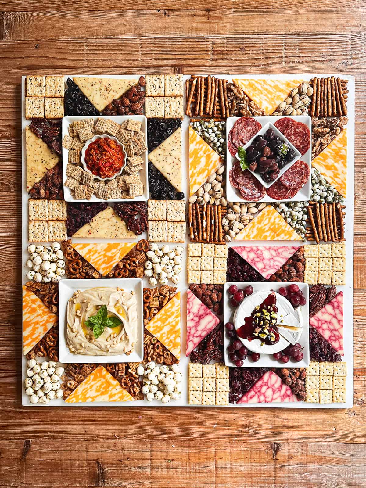 Americana charcuterie board with cheese, meats, nuts, etc. arranged in a patchwork quilt pattern on a wooden table.