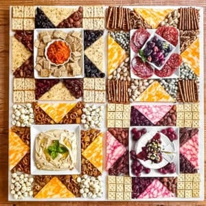 Americana charcuterie board with cheese, meats, nuts, etc. arranged in a patchwork quilt pattern on a wooden table.