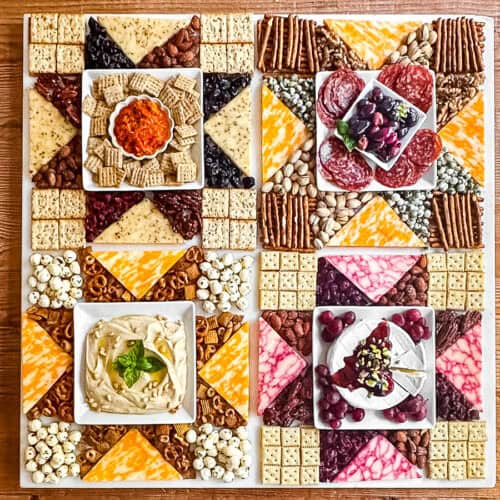 Americana charcuterie board with cheese, meats, nuts, etc. arranged in a patchwork quilt pattern on a wooden table.