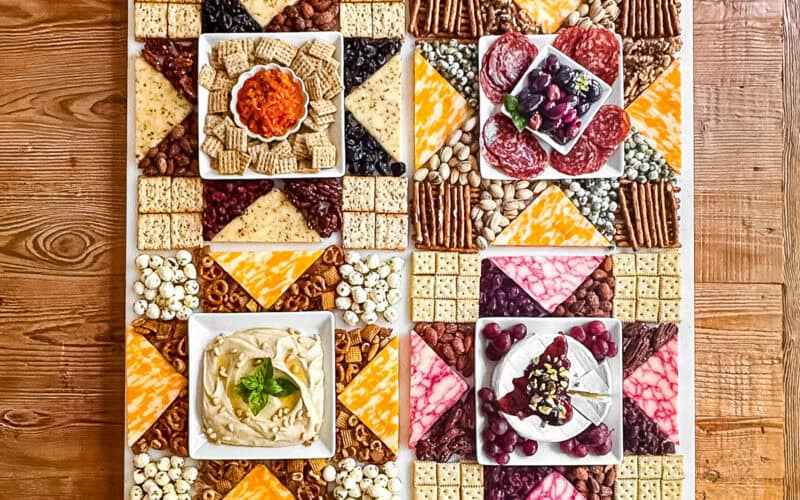 Americana charcuterie board with cheese, meats, nuts, etc. arranged in a patchwork quilt pattern on a wooden table.