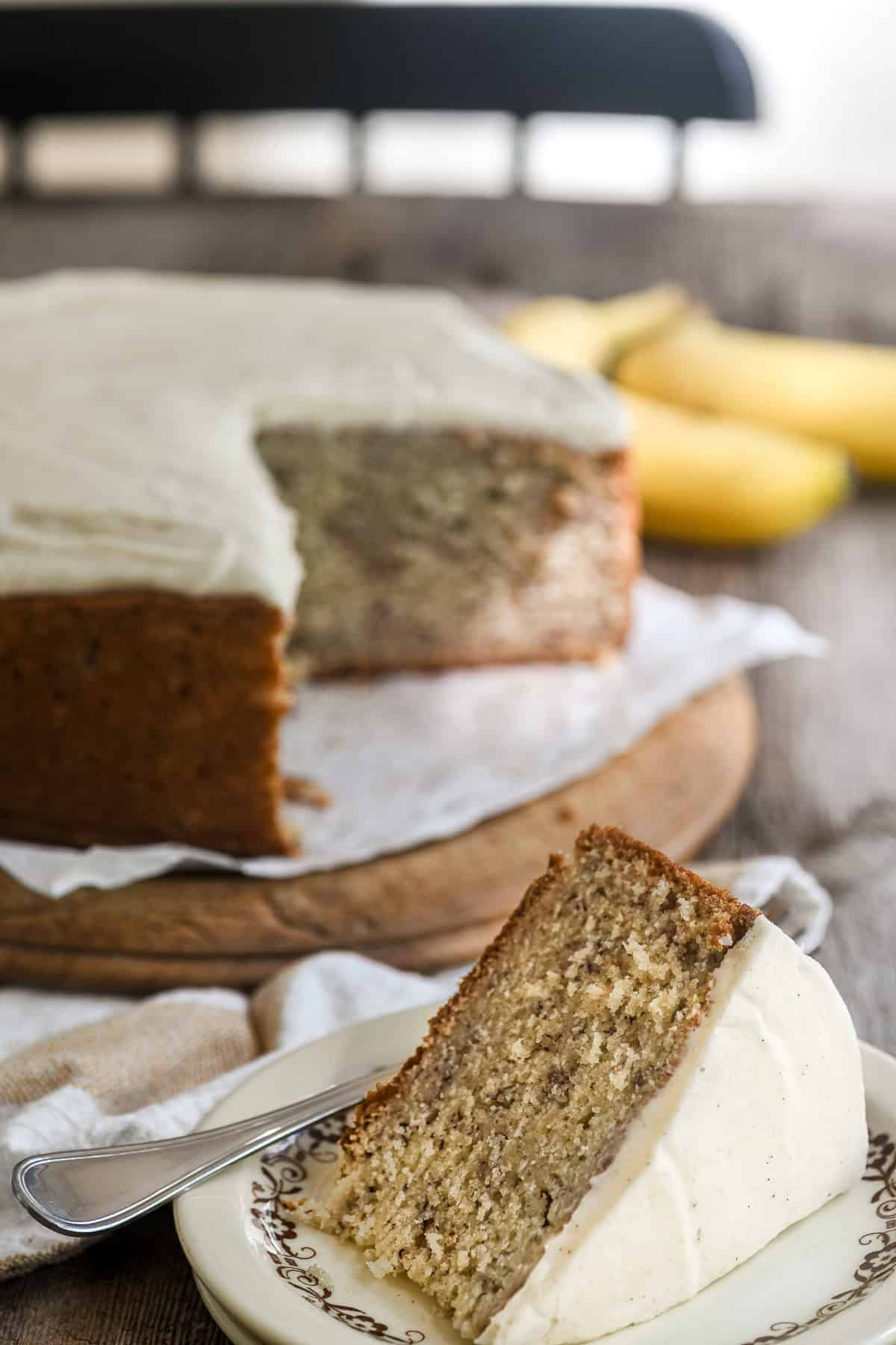 Kitchen table with banana cake topped with spiced mascarpone frosting, and bananas in the background.