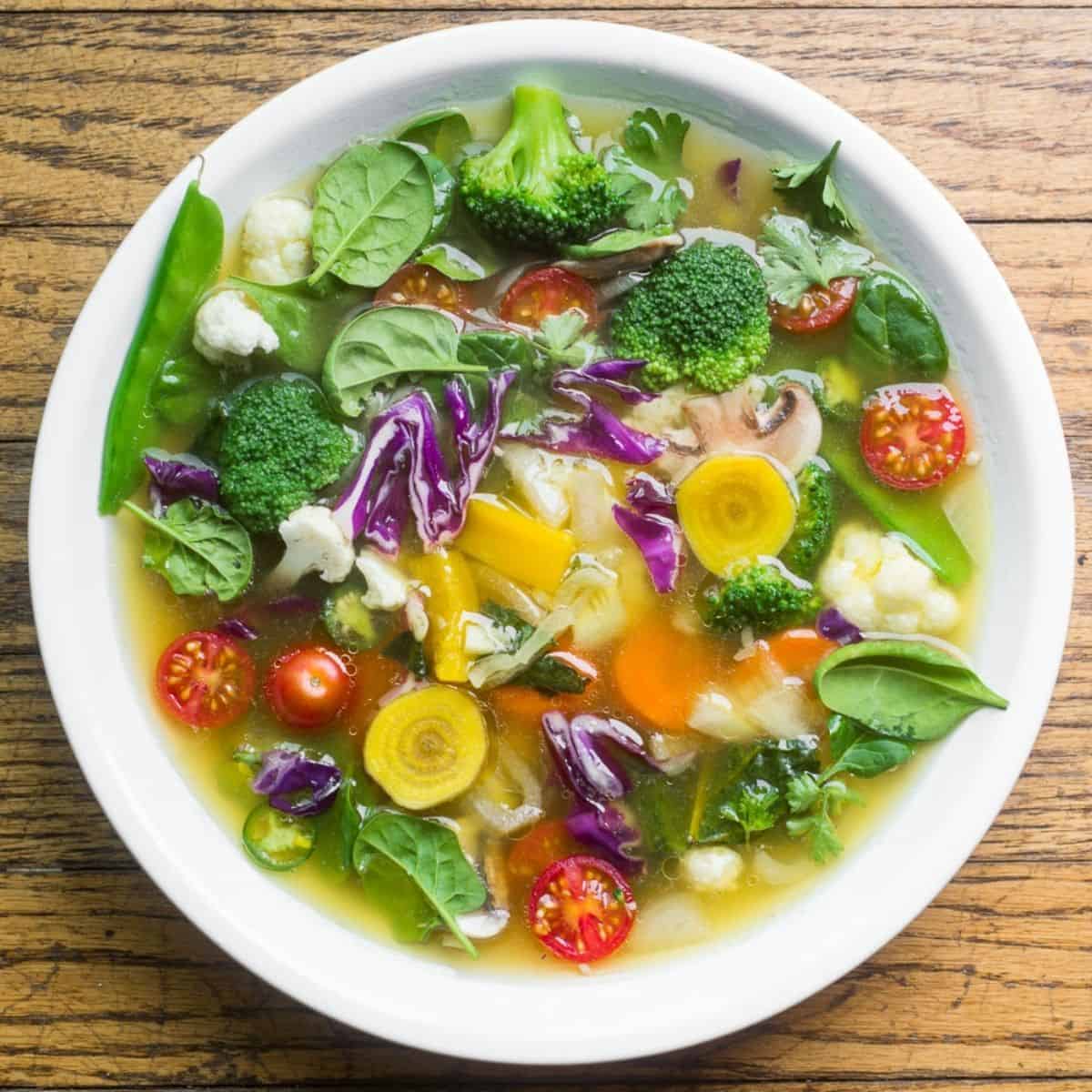 A white bowl of brothy vegetable soup on a wooden table.